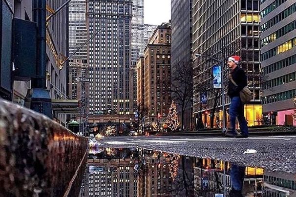 Photograph of man crossing midtown street on East Side while wearing Santa hat with reflection in puddle